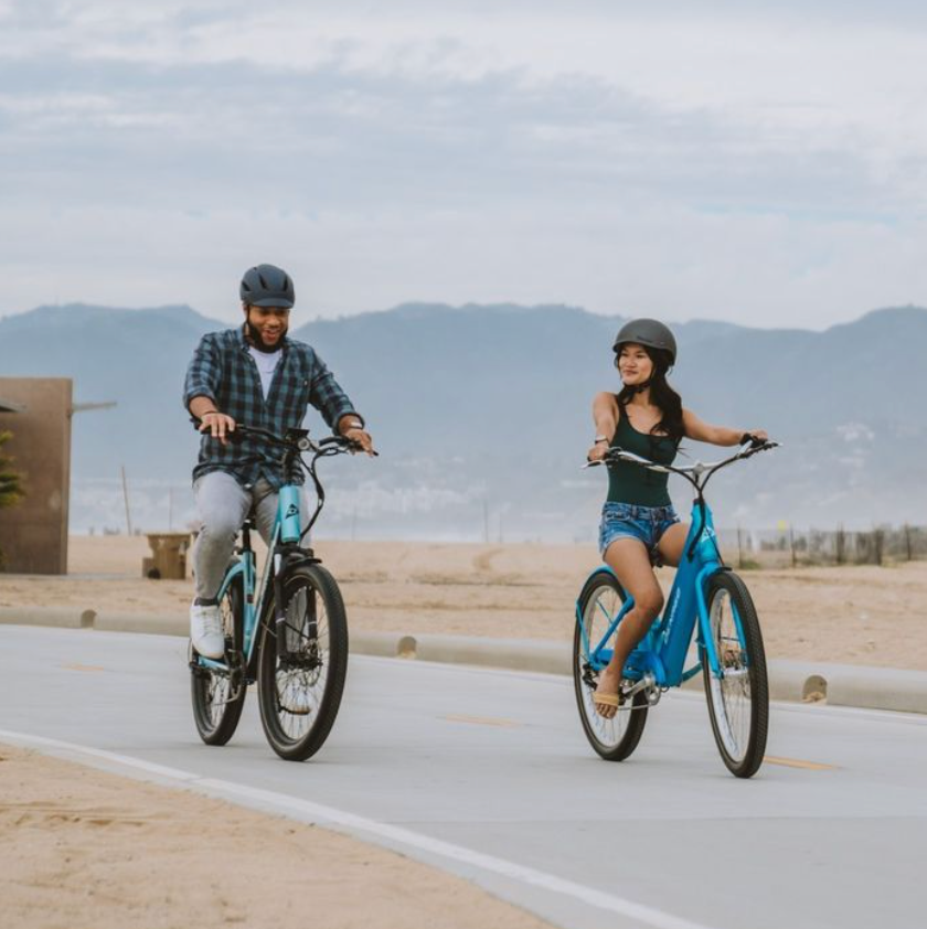 Couple riding bikes along the beach front with her on a teal Denago Cruiser 1.