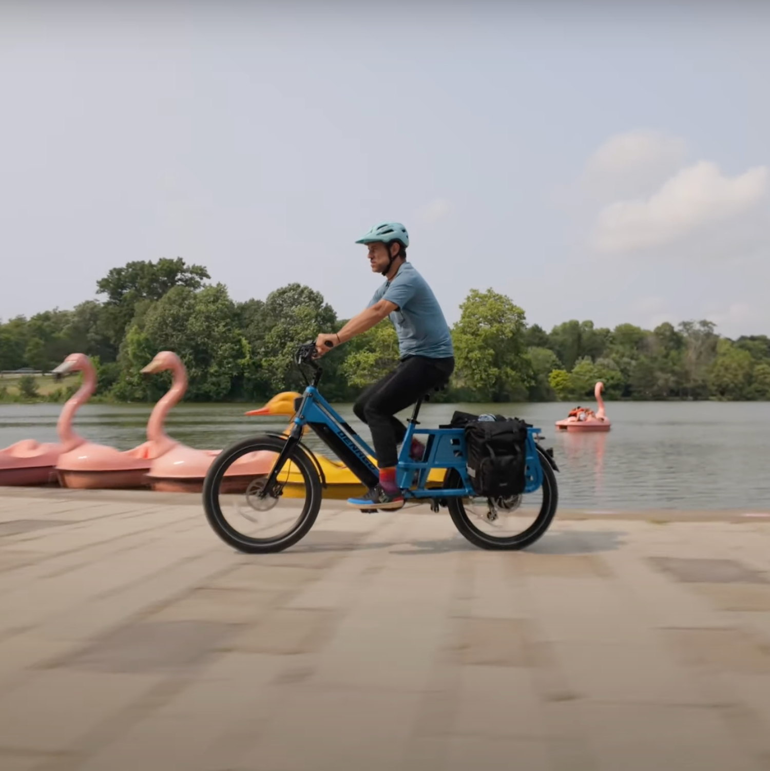 Guy riding by a lake on his Denago Cargo LT Electric Bike with flamingo and duck boats in behind him.