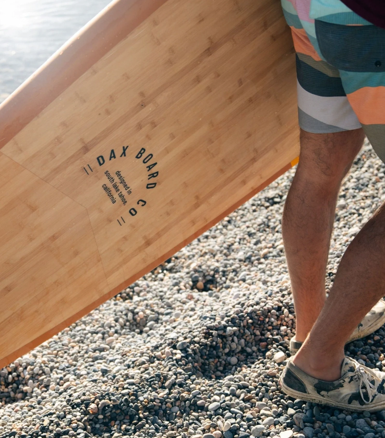 Guy carrying wood paddle board on rocky beach.