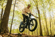 A man riding a Monarc Marker High-Step electric bike in Taconite with fat tires over a dirt trail in a sunlit forest with tall trees and green-yellow foliage.