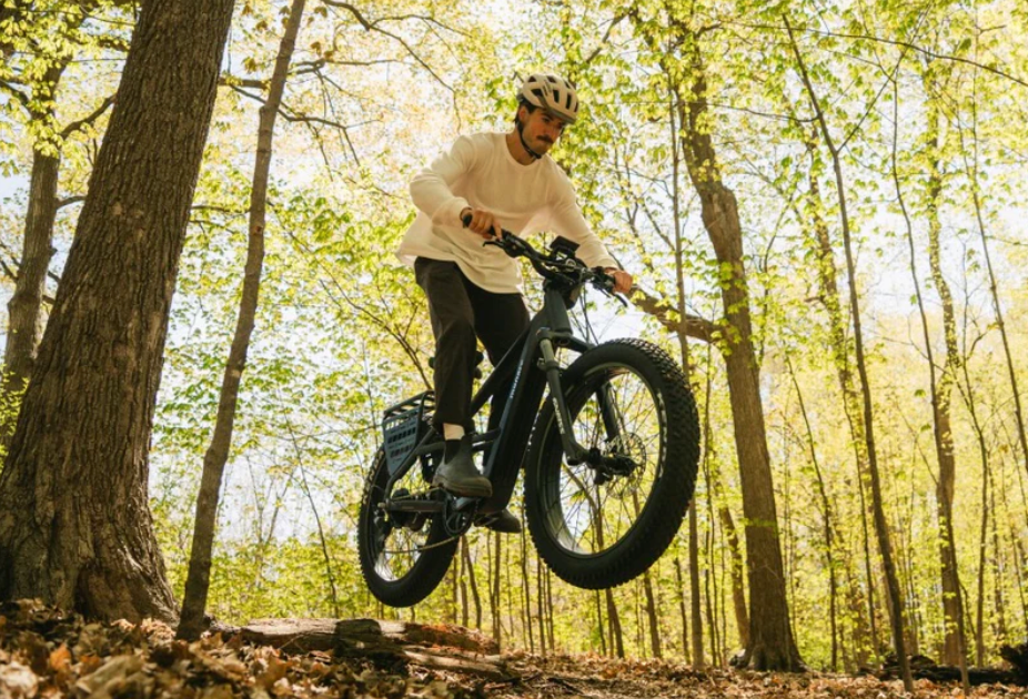 A man riding a Monarc Marker High-Step electric bike in Taconite with fat tires over a dirt trail in a sunlit forest with tall trees and green-yellow foliage.