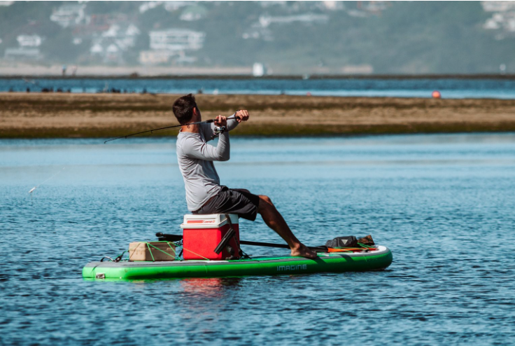 Guy fishing out on the water on his Imagine Surf Wizard Paddle Board sitting on a cooler.