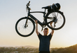 A man outdoors at sunset holding a matte black electric bike by Urtopia called the Carbon Classic above his head with both hands. The bike has a rear pannier bag and is shown against a warm sky with hills in the background.