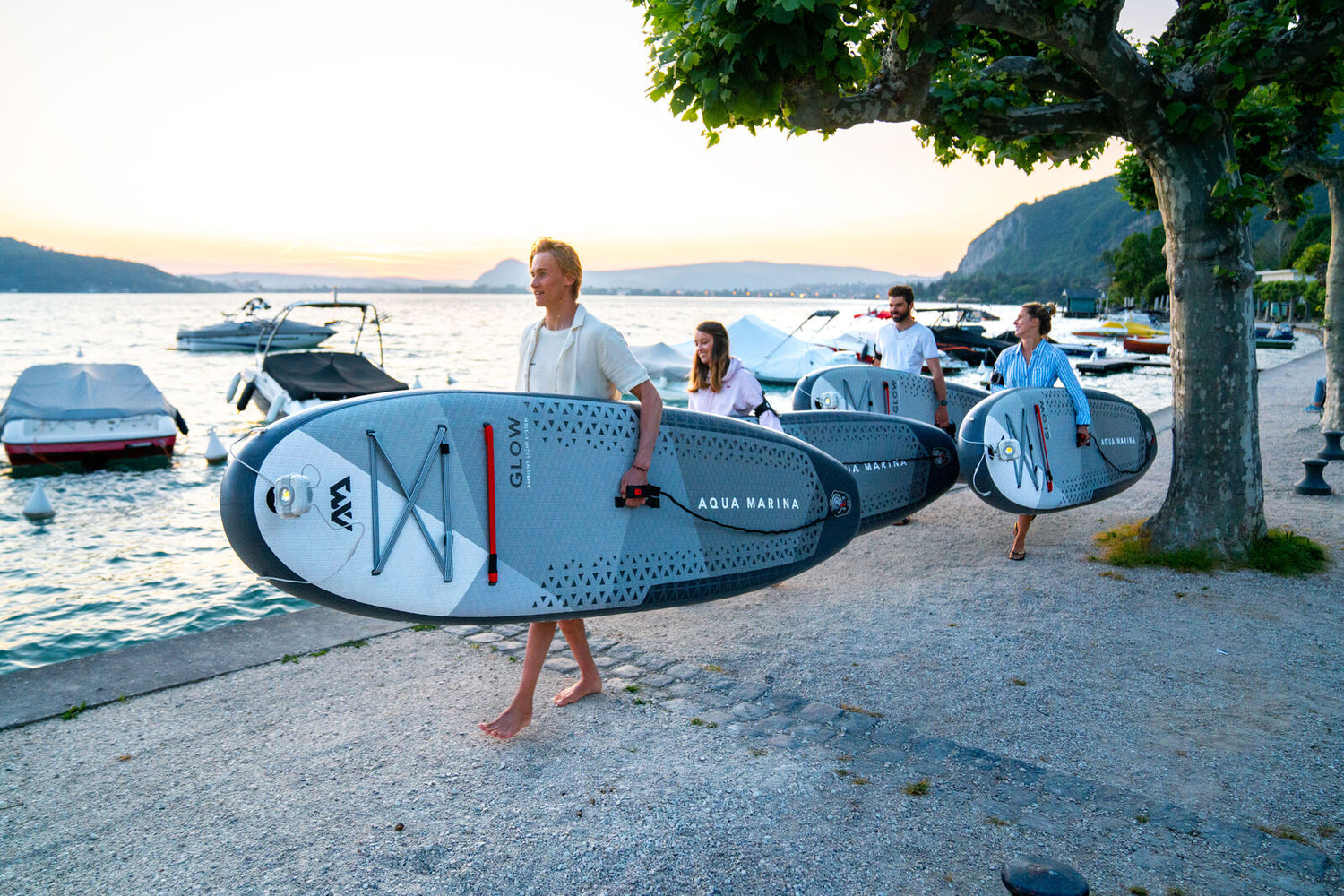 Group of people walking down the beach carrying their Aqua Marina Glow Inflatable Stand Up Paddle Boards with one hand.