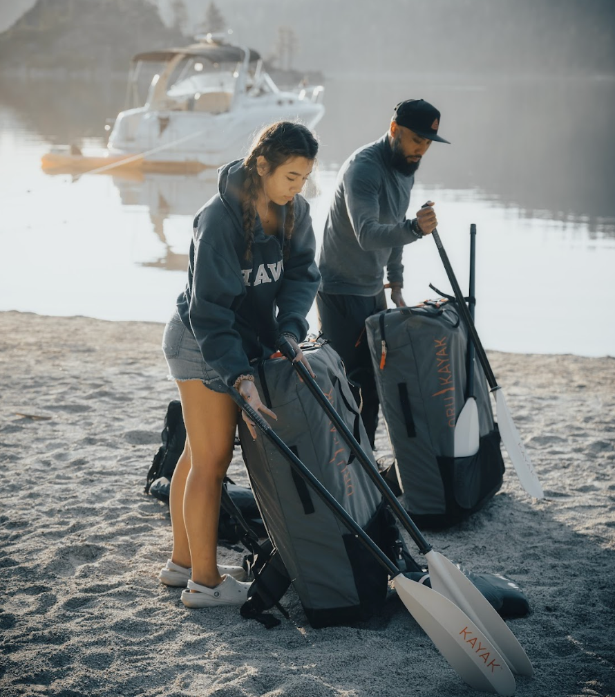 A couple each with their own Oru Kayak Coast XT in its backpack with paddles on a beach.