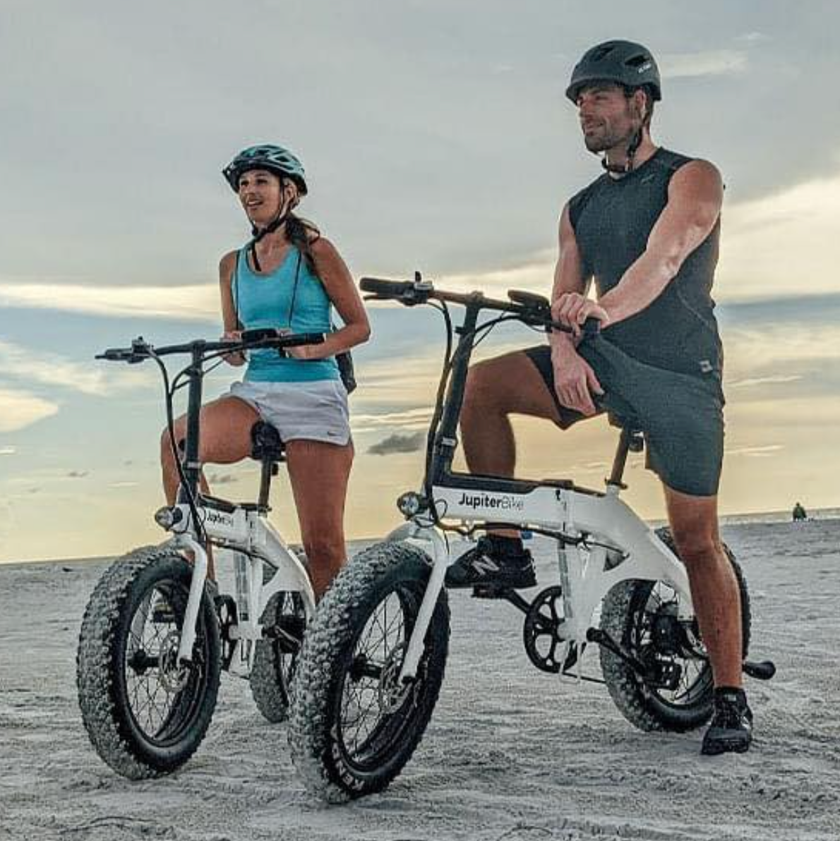 Couple riding two Jupiter Defiant Folding Electric Bikes on the beach with helmets on.