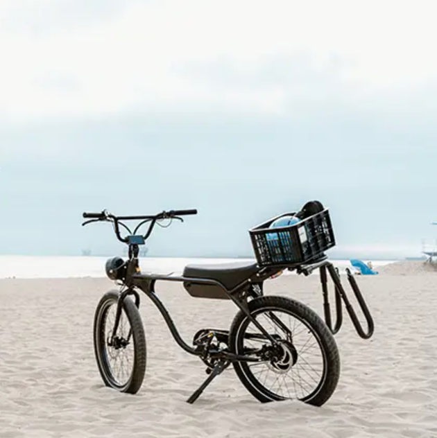 Model J from Electric Bike Company on a beach with surfboard rack and basket.