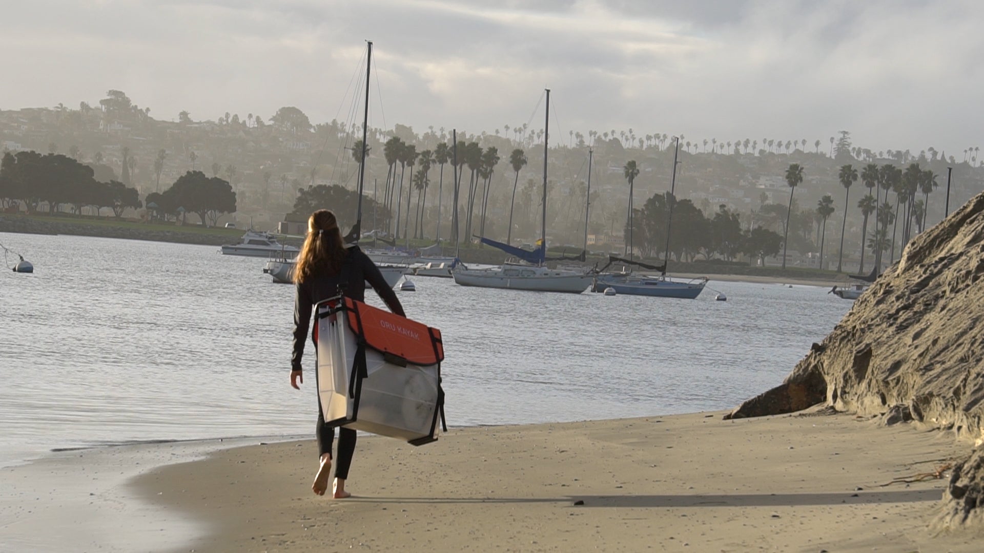 Girl walking on beach with foldable kayak over shoulder.