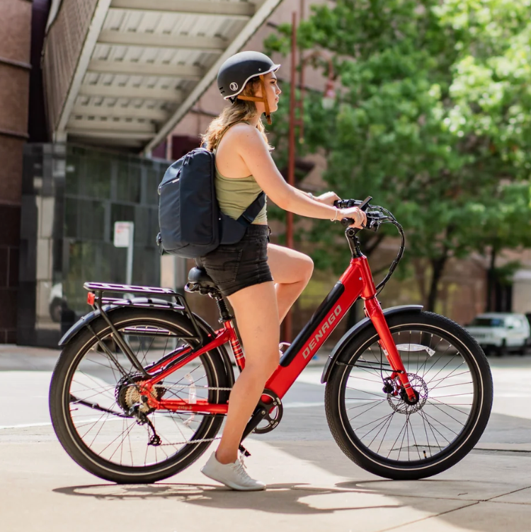 Girl with a backpack sitting on a red Denago City 3 Electric Bike with a building and trees behind her.