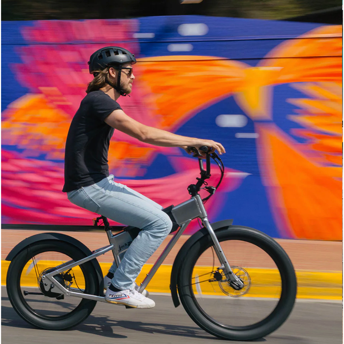Person riding an JackRabbit MG Renegade silver electric bike with a colorful mural in the background.