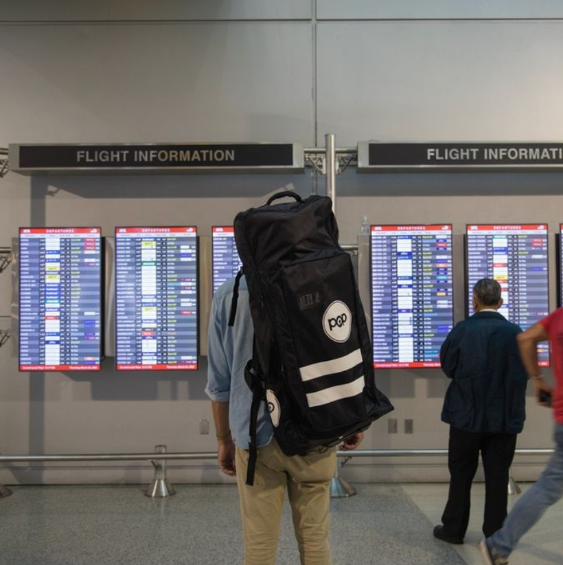 Guy standing looking at flight information at the airport with his POP Board Co. Royal Hawaiian SUP in the carrying bag.
