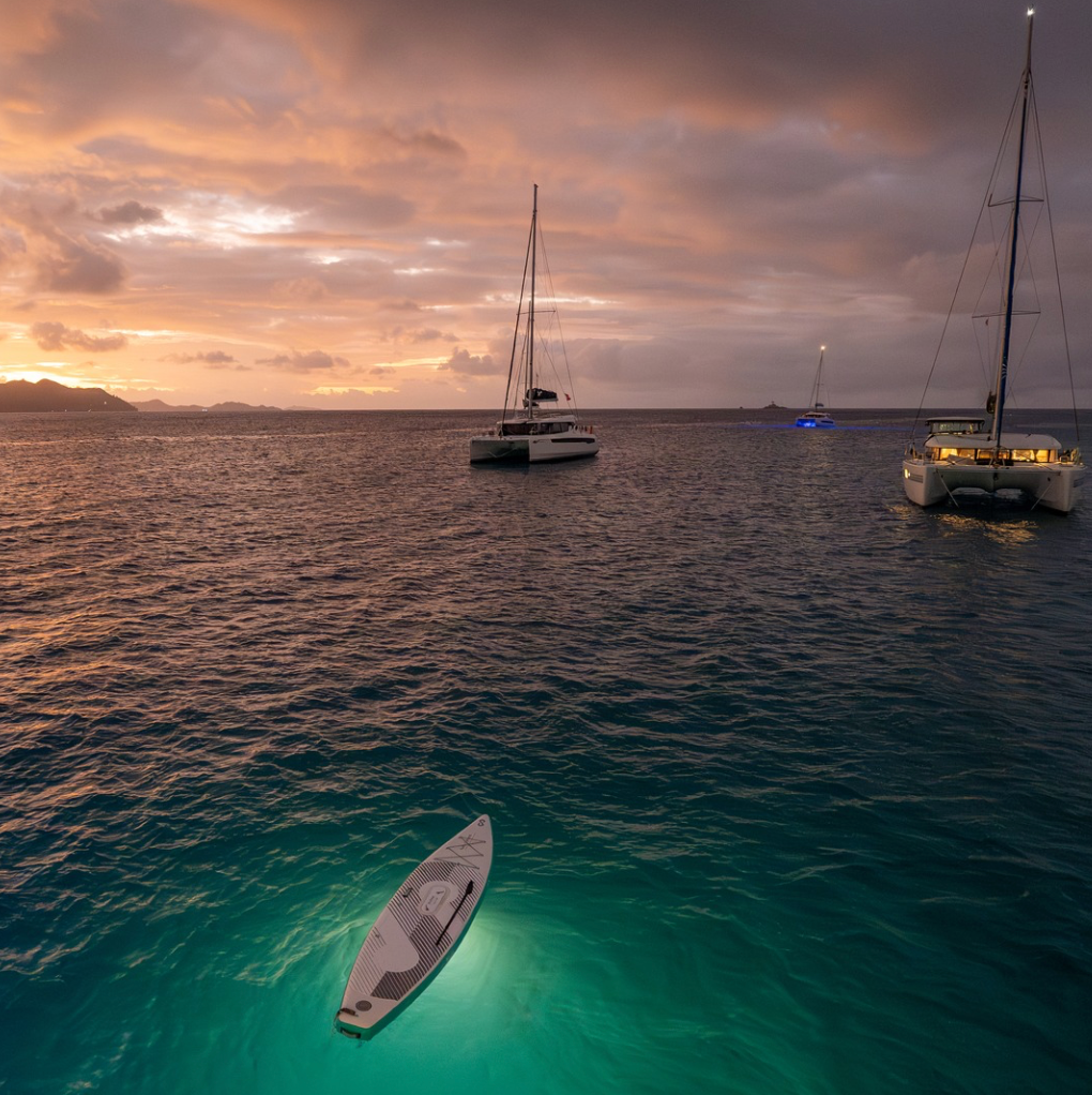 A SipaBoards Drive Neo Silver Inflatable SUP lit up on the water near sail boats at night.