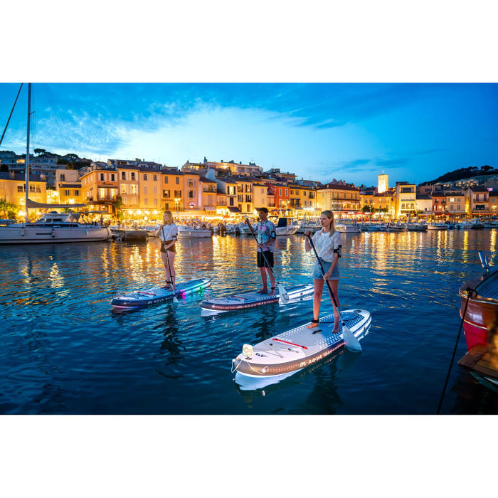 Three Aqua Marina Glow SUPs paddle boarders at night in marina with lights on. 