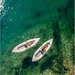 Two people in separate Aqua Marina Halve Kayaks floating in shallow water with paddles across their laps.