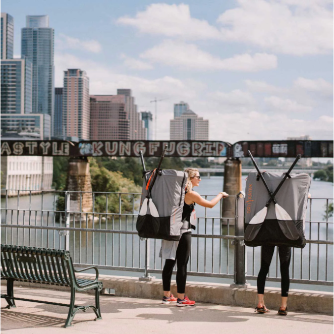 Two people standing on bridge over water with Oru Bay ST Kayak's in carrying cases on their backs.