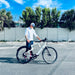Man standing behind a Urtopia Carbon 1 Pro eBike in a parking lot with trees and a fence in the background.