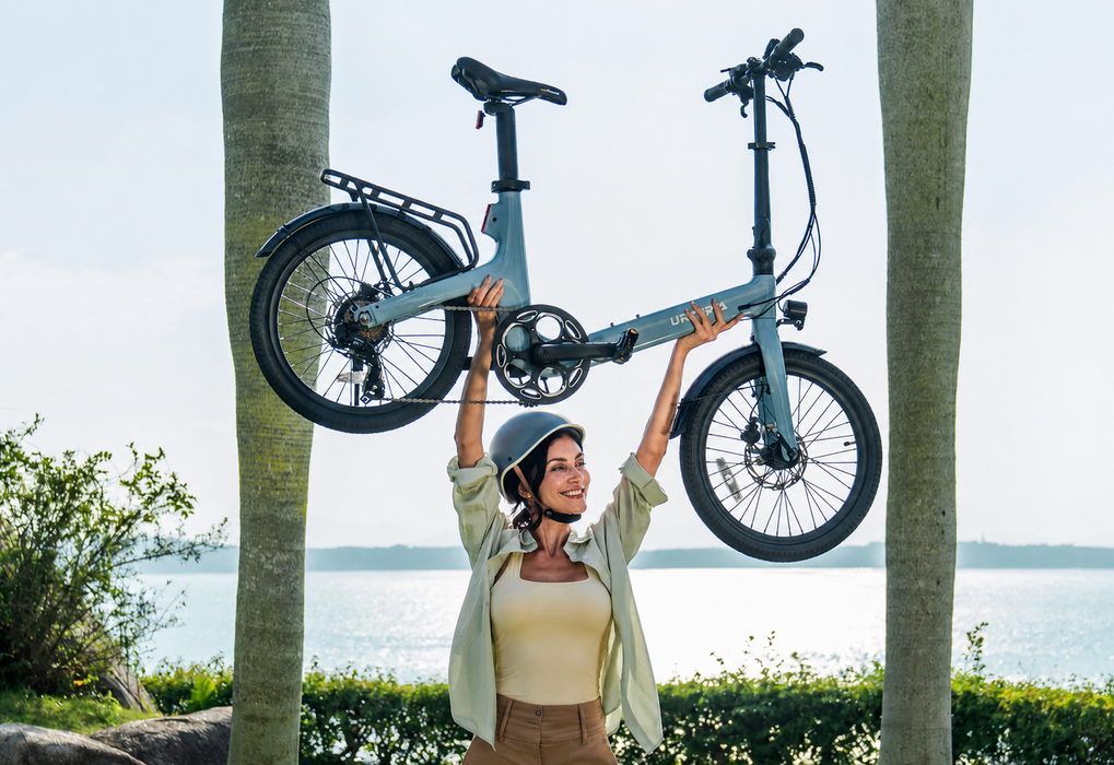 A woman in a helmet and light green shirt holding up an Urtopia Carbon Fold ST in Glacier Blue between two palm trees near a waterfront.